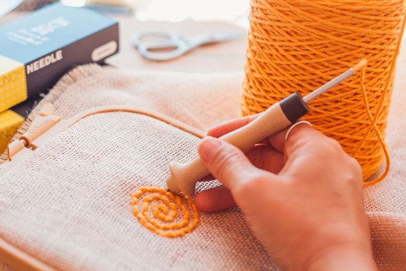 Pu&ograve; includere: Una mano utilizza uno strumento di legno per ricamare un motivo a spirale giallo su un tessuto beige teso in un telaio da ricamo. Un rocchetto di filo giallo si trova nelle vicinanze.