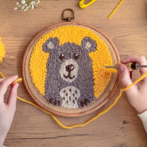 Puede incluir: Una persona está usando una aguja e hilo para crear un bordado de punto de aguja de un oso sobre un fondo amarillo. El bordado está en un aro de madera.