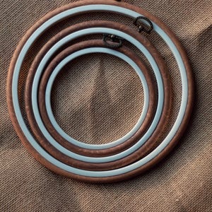 Könnte beinhalten: Drei hölzerne Stickrahmen mit weißen Innenringen, ineinander verschachtelt auf einem braunen Jutestoff-Hintergrund.