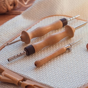 Pu&ograve; includere: Tre aghi da ricamo in legno con punte in metallo e un'asola in legno con la scritta "Linen" su di essa. Gli aghi sono su una superficie di tessuto beige.