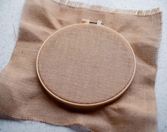 May include: A brown embroidery hoop with a beige fabric stretched across the frame. The hoop is lying on a piece of burlap fabric.