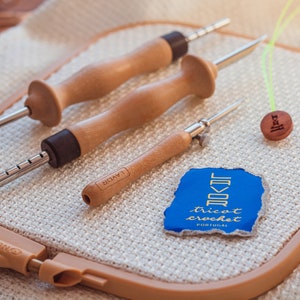 Pu&ograve; includere: Tre strumenti per uncinetto in legno con punte metalliche, un bottone in legno con la parola "fibra" incisa su di esso e un'etichetta blu e dorata con il testo "Tricot Crochet Portugal" su un cerchio da uncinetto beige.