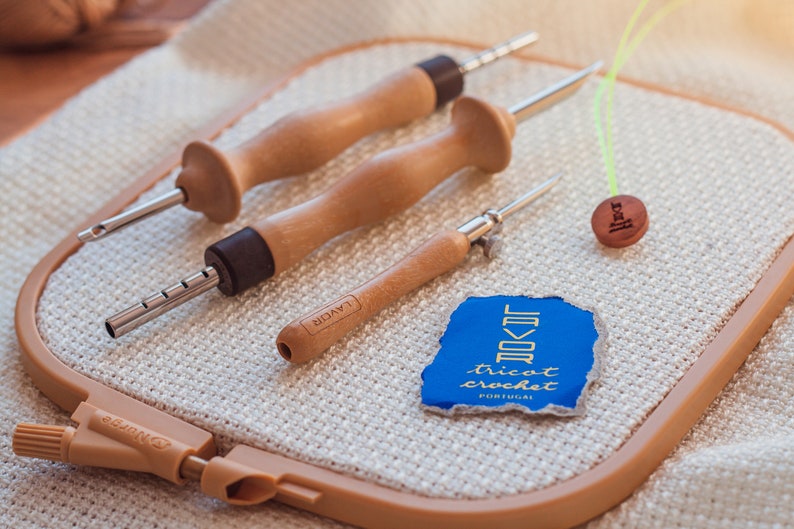 Pu&ograve; includere: Tre strumenti per uncinetto in legno con punte metalliche, un bottone in legno con la parola "fibra" incisa su di esso e un'etichetta blu e dorata con il testo "Tricot Crochet Portugal" su un cerchio da uncinetto beige.