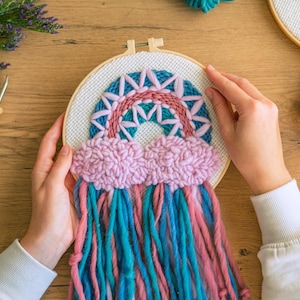 May include: A close-up of someone working on a rainbow-themed embroidery project. The embroidery hoop features a colourful rainbow with pink clouds and blue and pink yarn tassels.