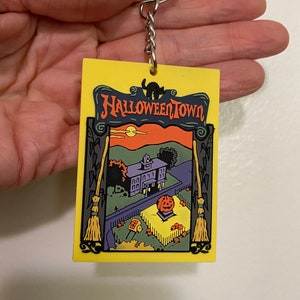 May include: A yellow Halloween Town keychain with a black cat, a house, and a pumpkin. The keychain is made of rubber and has a silver key ring.