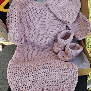 May include: A handmade, light mauve crochet baby set, including a gown, hat, and booties. The set is displayed in an open cardboard box. The crochet work has a textured appearance, with a consistent stitch pattern throughout the pieces. The set is designed for a newborn.