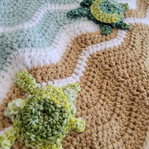 May include: Two crocheted green and white turtles on a crocheted tan, white, and blue blanket. The blanket has a wave pattern.