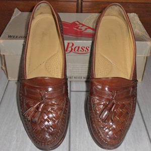 May include: A pair of brown leather tassel loafers with a woven pattern. The shoes are sitting on a wooden surface and have the word "Bass" printed on the box.