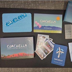 May include: A collection of Coachella music festival merchandise, including a welcome guide, memory book, postcards, and a box. The items feature the Coachella logo and desert landscape imagery, in shades of blue, pink, and orange.