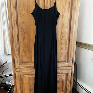 May include: A black spaghetti strap maxi dress hanging on a wooden hanger in front of a wooden cabinet.
