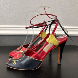 May include: A pair of vintage high-heeled sandals. The open-toe shoes feature red leather uppers with black and gold accents. The heels are black, and the ankle straps are red and black.