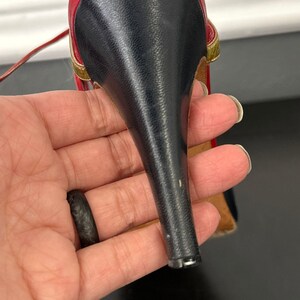 May include: A close-up of a high-heeled shoe. The shoe features a black heel, a red and gold strap, and a black upper. The shoe is held in a hand.