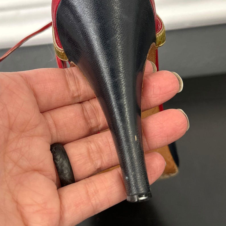 May include: A close-up of a high-heeled shoe. The shoe features a black heel, a red and gold strap, and a black upper. The shoe is held in a hand.