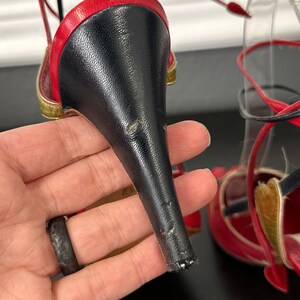 May include: Red and black high-heeled sandal with a stiletto heel. The sandal features a red upper with black accents and gold trim. The heel is black. The sandal has a strappy design.