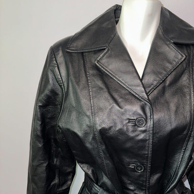 K&ouml;nnte beinhalten: Schwarze Lederjacke mit Reverskragen und Knopfverschluss. Die Jacke hat einen G&uuml;rtel und wird auf einer Schaufensterpuppe pr&auml;sentiert. Das Leder scheint glatt und leicht gl&auml;nzend zu sein.
