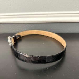 May include: A black belt with a textured, crocodile-like pattern. The belt has a silver-colored buckle with a decorative design. The inside of the belt is a light tan color.