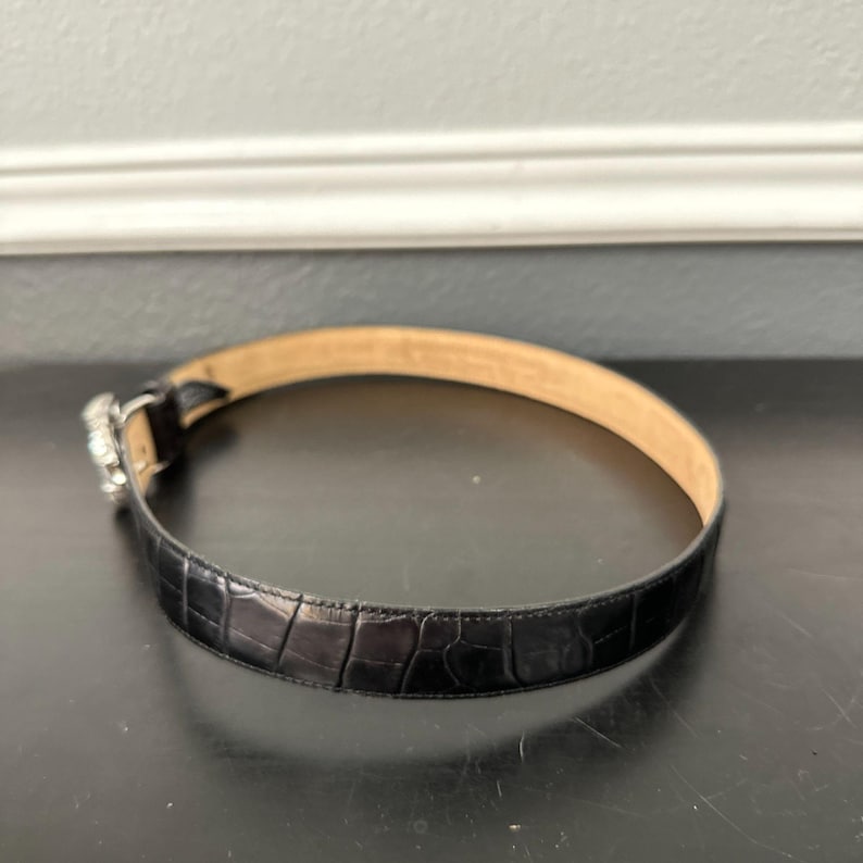 May include: A black belt with a textured, crocodile-like pattern. The belt has a silver-colored buckle with a decorative design. The inside of the belt is a light tan color.