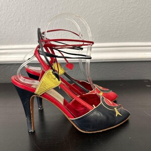 May include: A pair of high-heeled sandals with a black, red, and gold color scheme. The sandals feature a pointed toe, ankle straps, and a stiletto heel. The heel height is approximately 3 inches.