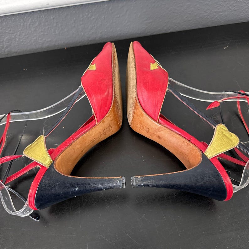 May include: Pair of red and black high-heeled shoes with a unique design. The shoes feature red leather uppers with gold accents, black heels, and clear straps. The heels are approximately 3 inches (7.6 cm) high.