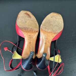 May include: A pair of vintage women's heels with red, black, and gold accents. The shoes have a heart-shaped detail on the ankle strap and a small heel. The soles are worn, and the size is 37.