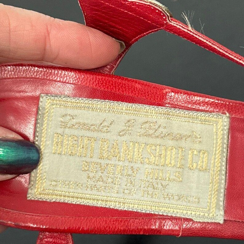 May include: Close-up of a red leather sandal with a gold buckle. A rectangular label with gold text reads "RIGHT BANK SHOE CO. BEVERLY HILLS MADE IN ITALY". The sandal is likely vintage.