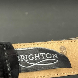 May include: Close-up of a black fabric label with the word "BRIGHTON" in white script. The label is sewn onto a tan leather strap, with a black background.
