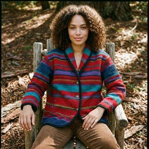 Puede incluir: Una chaqueta a rayas multicolores con ribete negro y dos botones. La chaqueta presenta rayas horizontales en rojo, azul, morado y turquesa. La persona lleva pantalones de pana marrones y está sentada en una silla de madera.