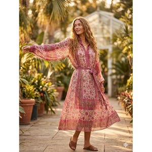 May include: A long-sleeved, flowing dress with a floral pattern in shades of pink and cream. The dress features a V-neck, a belted waist, and a midi-length skirt. The sleeves are long and billowy. The dress is worn with brown sandals.