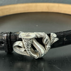 May include: Black leather belt with a silver-toned buckle. The buckle features an ornate design with a stylized letter "S" and decorative scrollwork. The belt has a textured, crocodile-like pattern.