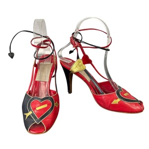 May include: A pair of red leather open-toe heels with black and gold heart accents. The heels have a pointed toe, a thin ankle strap, and a stiletto heel. The design includes a heart pierced by an arrow.