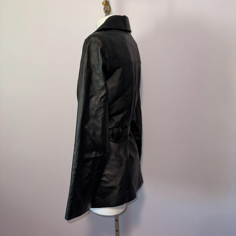 K&ouml;nnte beinhalten: Schwarze Lederjacke mit Reverskragen und G&uuml;rteldetail in der Taille. Die Jacke wird auf einer wei&szlig;en Schaufensterpuppe pr&auml;sentiert und hat ein klassisches, ma&szlig;geschneidertes Design. Das Leder scheint eine leicht strukturierte Oberfl&auml;che zu haben.