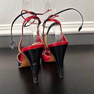 May include: A pair of high-heeled sandals with a black heel, red and gold accents, and black ankle straps. The open-toe shoes have a stiletto heel and are displayed on clear plastic forms.