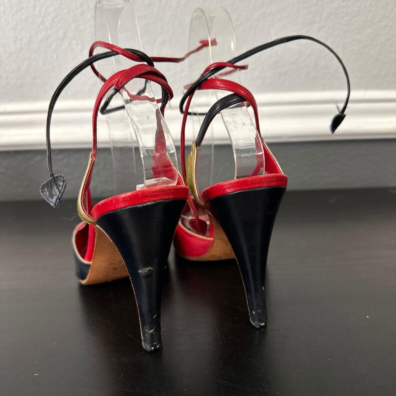 May include: A pair of high-heeled sandals with a black heel, red and gold accents, and black ankle straps. The open-toe shoes have a stiletto heel and are displayed on clear plastic forms.