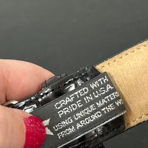 May include: Close-up of a black leather belt with a textured finish. A tag reads "Crafted with Pride in U.S.A. Using Unique Materials From Around the W". The belt is held by a person with red nail polish.