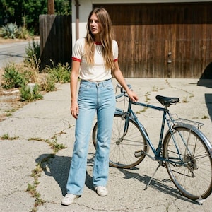 Può includere: Una donna è in piedi accanto a una bicicletta blu vintage. Indossa una maglietta color crema con rifiniture rosse, jeans azzurri e scarpe da ginnastica bianche. La bicicletta ha una sella nera e un cestino argentato.