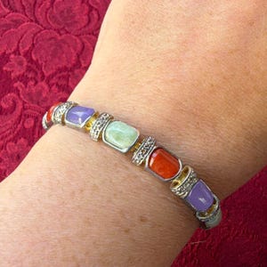 Op de afbeelding: Een schakelarmband met rechthoekige, glad gepolijste stenen in tinten lavendel, lichtgroen en oranje. Elke steen is gezet in een zilverkleurig metalen frame met kleine, heldere, sprankelende accenten langs de verbindingsschakels.