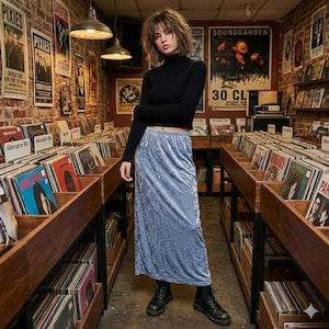 May include: A woman wearing a black turtleneck sweater and a long, light blue velvet skirt, paired with black boots. The setting is a record store, with shelves of vinyl records and vintage posters on the brick walls.