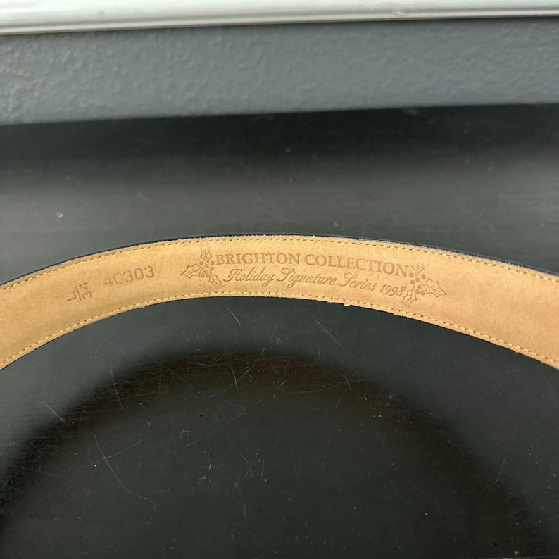 May include: A close-up of the inside of a black leather belt. The belt is stamped with the text "BRIGHTON COLLECTION Holiday Signature Series 1998". The size is 32, and the style number is 4C303.