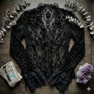Puede incluir: Blusa de encaje negro con mangas largas, cuello en V y cierre de lazo. La blusa presenta un estampado floral y ribete con volantes. Los puños están adornados con encaje y lazos de cinta. También se ven una baraja de cartas del tarot y cristales de amatista.