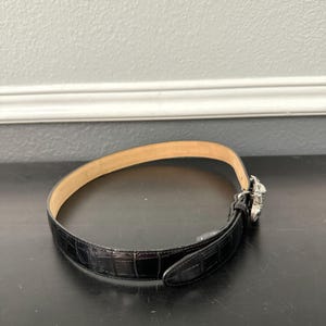 May include: Black leather belt with a crocodile pattern. The belt has a silver-colored buckle with a decorative design. The interior of the belt is a light tan color. The belt is on a black surface.