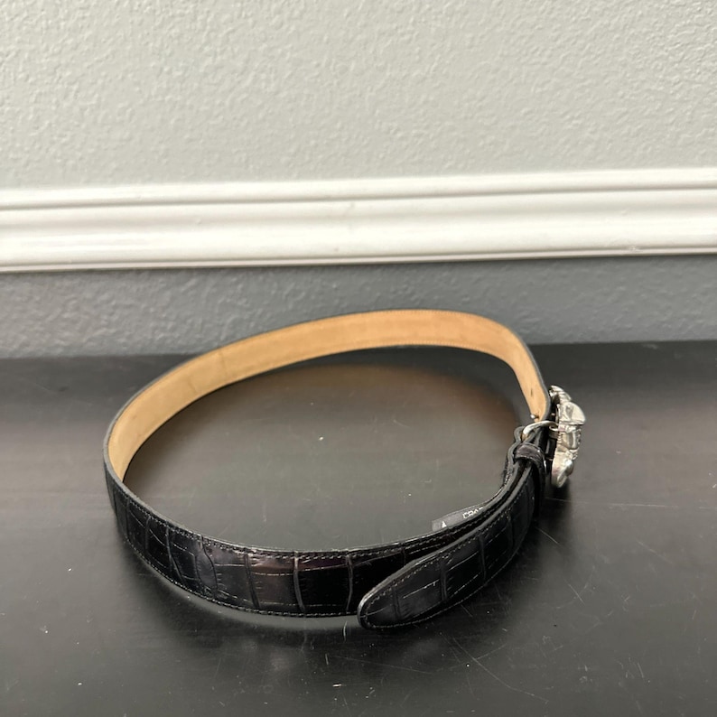 May include: Black leather belt with a crocodile pattern. The belt has a silver-colored buckle with a decorative design. The interior of the belt is a light tan color. The belt is on a black surface.