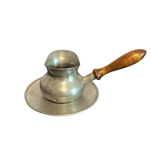 Op de afbeelding: Een kleine, zilverkleurige metalen koffiepot met een houten handvat. De pot staat op een bijpassende ronde schotel. Het handvat is lichtbruin van kleur. De pot heeft een gebogen tuit en een afgeronde bovenkant.