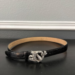 May include: Black leather belt with a silver-toned buckle featuring a musical horn design. The belt has a textured, crocodile-like pattern and a light brown interior. The buckle is the focal point, adding a decorative element to the accessory.