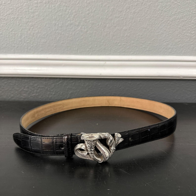 May include: Black leather belt with a silver-toned buckle featuring a musical horn design. The belt has a textured, crocodile-like pattern and a light brown interior. The buckle is the focal point, adding a decorative element to the accessory.