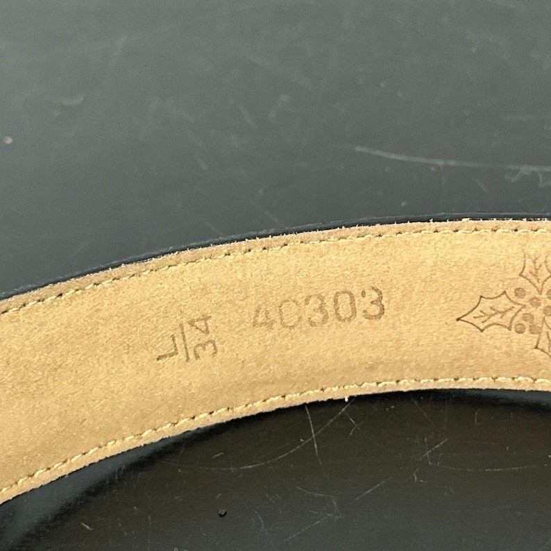 May include: Close-up of a black belt with a tan leather interior. The interior displays the size 34L, the number 40303, and a snowflake design. The belt is curved and appears to be made of leather.