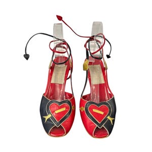 May include: A pair of red and black open-toe sandals with heart-shaped designs. The sandals feature gold arrow accents and tie-up ankle straps. The heels are approximately 3 inches high.
