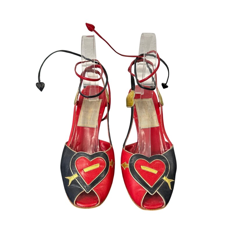 May include: A pair of red and black open-toe sandals with heart-shaped designs. The sandals feature gold arrow accents and tie-up ankle straps. The heels are approximately 3 inches high.