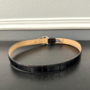 May include: Black leather belt with a crocodile pattern. The belt has a silver-colored buckle and a light brown interior. The belt is approximately 1 inch wide.