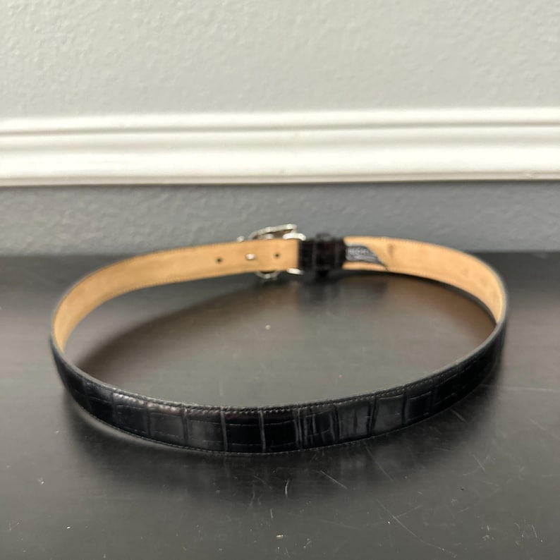 May include: Black leather belt with a crocodile pattern. The belt has a silver-colored buckle and a light brown interior. The belt is approximately 1 inch wide.