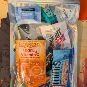 Może przedstawiać: Przezroczysta, opalizująca torebka strunowa wypełniona produktami do pielęgnacji i zdrowia w rozmiarze podróżnym. Zawiera płyn do demakijażu Neutrogena, miętówki Tic Tac, mieszankę witamin Emergen-C, chusteczkę Shout i batonik Mini's. Torebka jest zamknięta.